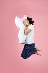 Young Asian woman in pajamas and an eye mask is jumping high and resting her head on a pillow on a pink background.