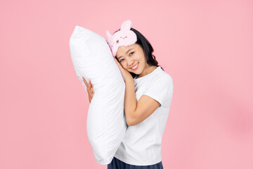 A young Asian woman in pajamas and an eye mask rests with her head on a pillow on a pink background.