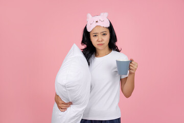 A sleepy, beautiful young Asian woman in pajamas and an eye mask holds a cup of hot coffee. She yawns against a pink background in a studio. A concept of waking up in the morning.