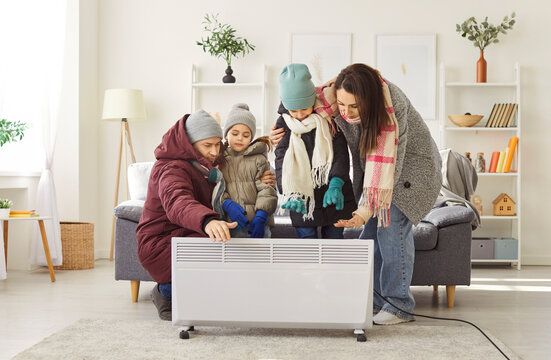 Young family wearing warm clothes standing together near home heater in cold room, trying to stay warm. Parents and children enduring chilly weather and dealing with heating issues in their house.