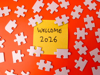 Fototapeta premium Yellow sticky note with the text WELCOME 2026 surrounded by white jigsaw puzzle pieces on an orange background