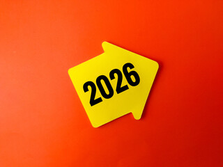 The image displays a yellow arrow-shaped sign with the number 2026 written in bold black font, set against an orange background