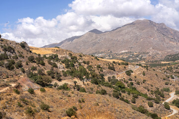 scenic landscape of the rugged Cretan mountains and valleys in Greece, captured on clear sunny day. Image features dramatic rocky peaks, rolling hills, scattered vegetation, and patches of farmland