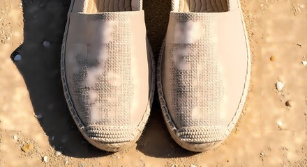 Fototapeta premium Pair of Beige Canvas Espadrille Shoes Resting on Sandy Beach with Sunlight Shadows