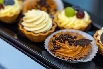 Close-up of assorted mini cream tarts. A detailed view of various mini dessert tarts with rich cream toppings, fruit garnishes, and chocolate decorations on a black tray.