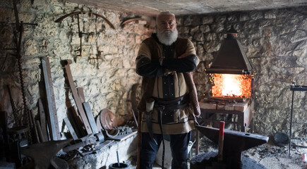 A Viking blacksmith with a white beard and braided hair works intently at a forge, hammering glowing metal on an anvil, with flames flickering in the background of a stone workshop.