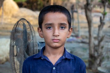 Serious Young Boy Standing Outdoors in Natural Light with a Rural Village Background