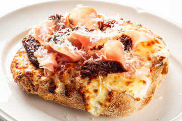 A delicious pizza or flatbread topped with melted mozzarella cheese, savory prosciutto, sundried tomatoes, and grated parmesan, on a white plate
