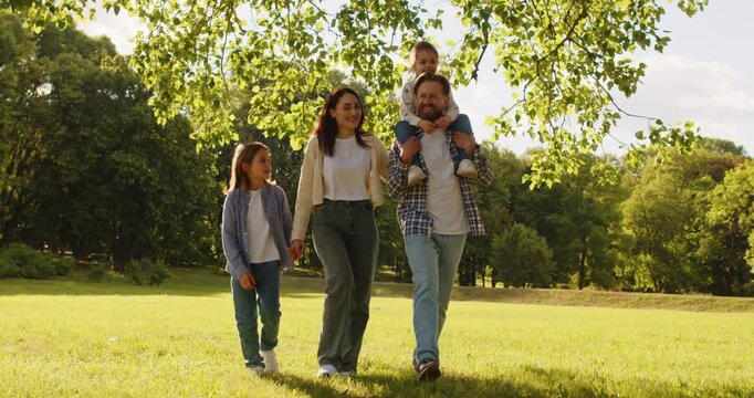 Summer walk of happy family in green park. Four people walking together on grass, parents and kids talking and enjoying nature at picnic. Dad carrying daughter on shoulders, mom holding hand of son