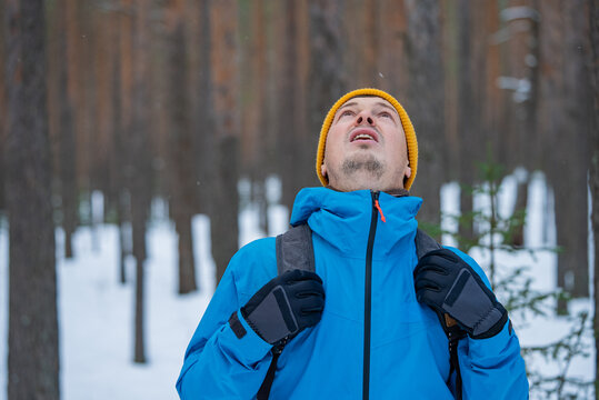 Man wearing a yellow beanie and blue winter jacket, with a backpack, looking up while standing in a snowy forest, appreciating the serene winter landscape and falling snow