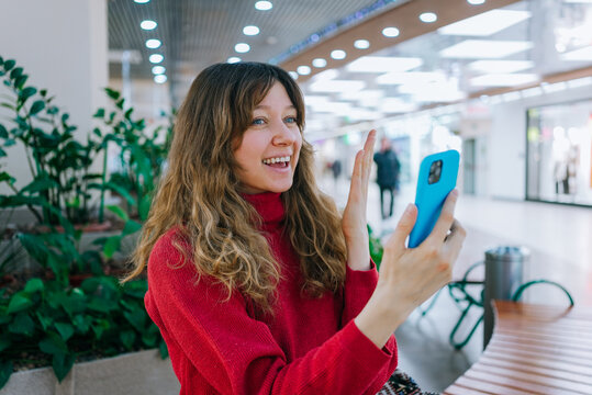 Woman in red sweater communicating remotely, holding phone and waving to camera during video call in a brightly lit shopping center, sharing her day with family or friends