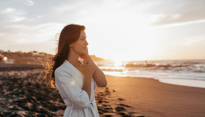 Serene woman in plush robe embraces golden sunset glow on tranquil beach, finding peaceful escape and rejuvenation by the ocean.