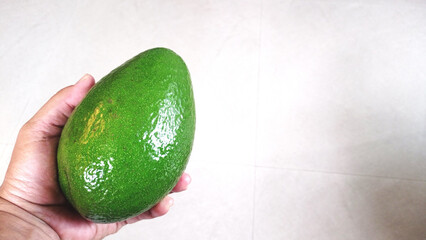 Whole green avocado held over neutral tiled background. Healthy eating, dieting, fresh fruit, Mexican food, guacamole ingredient