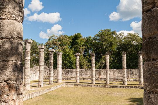 Exploring ancient Mayan city ruins, stone columns rise from the grassy ground, showcasing the historical architecture and archaeological site under a vibrant blue sky in Chichen Itza, Mexico - Powered by Adobe