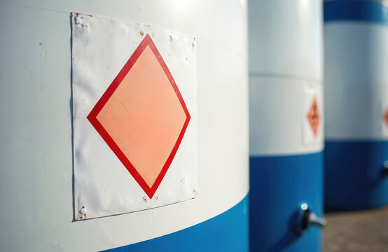 Industrial barrels with hazard symbols are lined up outdoors. White and blue drums show orange diamond labels. These containers store hazardous liquids or chemicals.