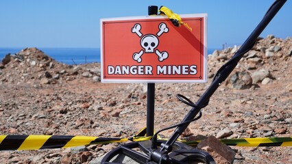 Metal detector resting next to a DANGER MINES sign and hazard tape on a rocky, desolate landscape,...