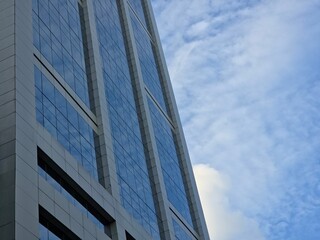 Low-angle view of the glass and steel facade of a modern skyscraper, reflecting the blue, slightly cloudy sky.