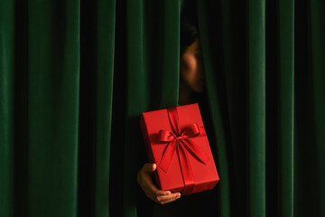 Mysterious person hidden behind dark green velvet curtain holding bright red gift box with satin bow, dramatic Christmas surprise and secret present concept, generative AI