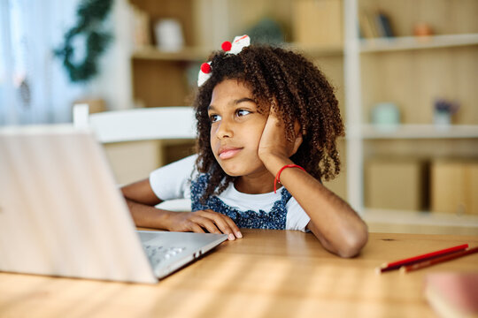Portrait of teenage black girl using laptop computer and thinking and day dreaming looking through window at home. Teenage girl attending to online school class worried and tired with headache