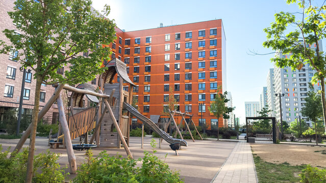 Modern urban playground with wooden slides, swings, and climbing structures surrounded by colorful apartment buildings and greenery, representing family lifestyle and city comfort concepts