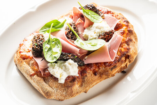 A gourmet Italian flatbread featuring mortadella, fresh ricotta cheese, and wild morel mushrooms, garnished with basil leaves on a white plate
