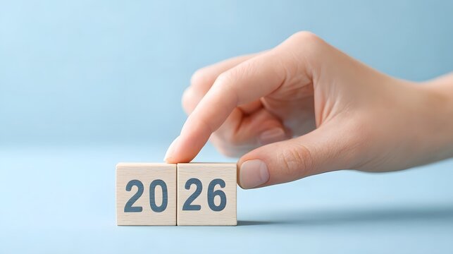 Close up of a hand about to touch wooden blocks with the numbers 20 and 26 symbolizing the upcoming year 2026 against a clean blue background