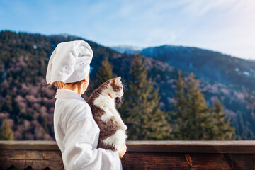 Obraz premium Woman holding cat on hotel balcony enjoying winter mountain landscape wearing bathrobe. Travel with pet