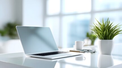 A sleek silver laptop rests on a polished white desk complemented by a white coffee mug a green potted plant and a notebook illuminated by abundant natural light from large in a minimalist workspace