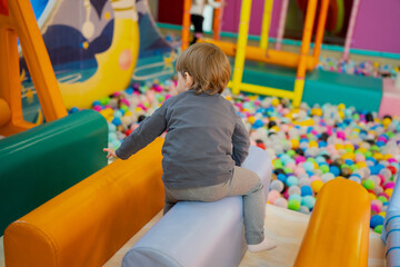 Child playing on cushioned rollers inside a bright indoor play area, with a large ball pit and soft...