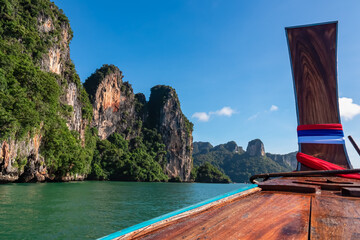 The prow of a traditional Thai long-tail boat sails through emerald Andaman Sea, offering stunning scenic view of majestic limestone cliffs and lush jungle of Railay Bay under clear blue sky in Krabi