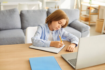 Concentrated schoolboy writing homework in a notebook while engaging in remote learning, sitting at desk at home, demonstrating focus on education, thinking, and maintaining school assignments.