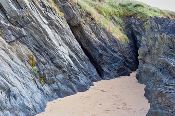 A landscape photo depicts a sandy cove with steep, layered rock formations on either side, partially covered with green foliage at the top in Polly Joke - Cornwall - UK