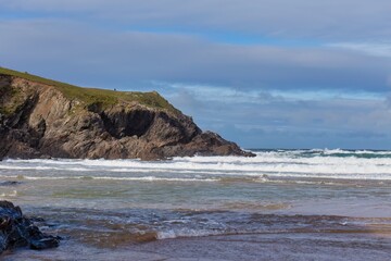 Coastal landscape photo. Waves roll onto a sandy beach, with a rocky headland in the background beneath a partly cloudy sky in Polly Joke - Cornwall - UK