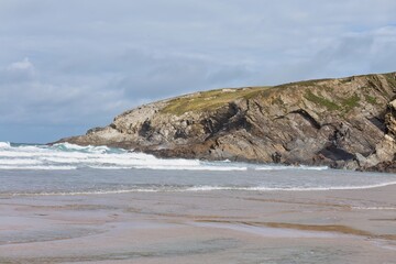A rocky headland meets a sandy beach. Waves break gently on the shore under a cloudy sky in Polly Joke - Cornwall - UK
