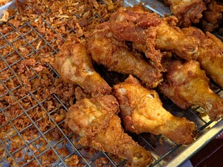 Close-up of a pile of golden brown, crispy fried chicken drumsticks displayed on a metal cooling rack next to a large heap of crispy fried shallots.