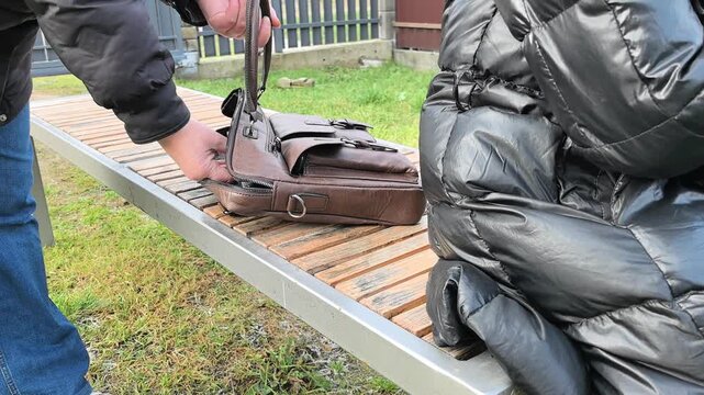 In a public area, an individual approaches a bag placed on a bench while another person sits unaware. The setting is outdoors, suggesting a possible theft of a laptop.