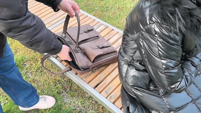 In a public area, an individual approaches a bag placed on a bench while another person sits unaware. The setting is outdoors, suggesting a possible theft of a laptop.