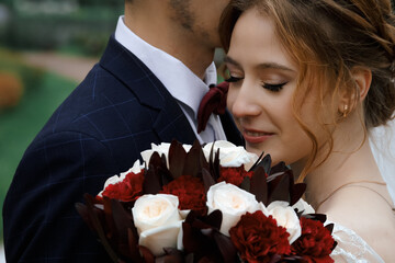 A tender wedding moment: a bride with closed eyes smiles softly, nestled against her groom in a navy suit. They hold a stunning bouquet of red and white roses, capturing romance and joy