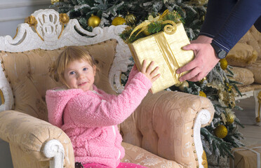 a little girl in a pink sweater is sitting in a chair and being handed a large box wrapped in Christmas paper