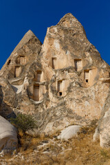 Cappadocia. T&uuml;rkiye. Sunlit, beige rock formations with carved windows and doorways against a deep blue sky, surrounded by dry grass and sparse shrubs.