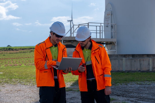 Wind energy technicians inspect turbine site while reviewing data on a laptop during sunny afternoon