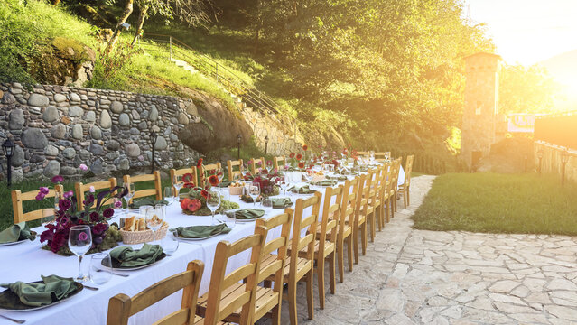 Long outdoor dining table beautifully set with flowers, fruits, and glassware in a scenic green garden near stone walls, symbolizing celebration, hospitality, and rustic elegance