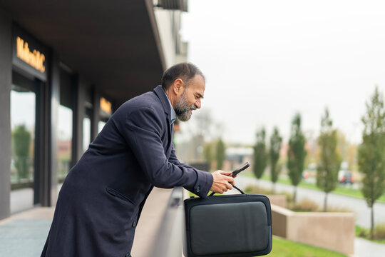 Mature businessman checking smartphone outdoors holding brief case