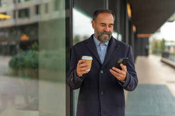 Mature businessman checking smartphone holding coffee in city