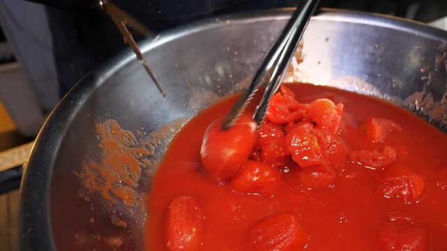 A cook wearing black gloves uses kitchen scissors and metal tongs to cut whole peeled tomatoes directly inside a large stainless steel bowl filled with tomato sauce.
