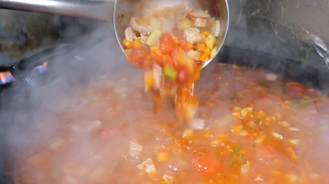 A gloved chef uses a large metal ladle to scoop and transfer steaming hot chicken and vegetable stew from a boiling pot on a stove into a stainless steel chafing dish or serving container in a commerc