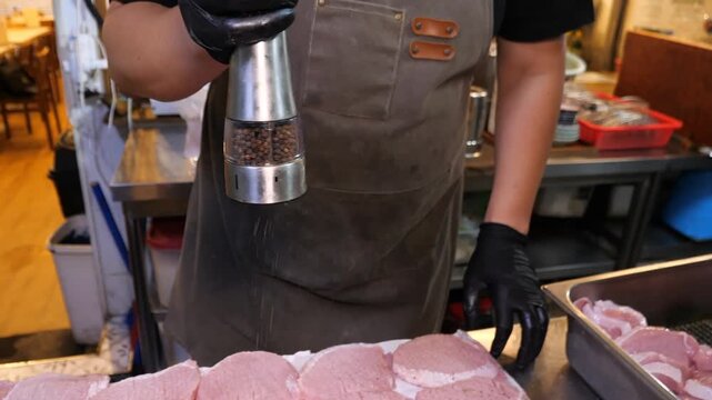 A gloved chef uses a metal shaker to sprinkle ground pepper onto raw, tenderized pork chops arranged on paper towels, then flips and places additional chops onto the board for seasoning in a kitchen.