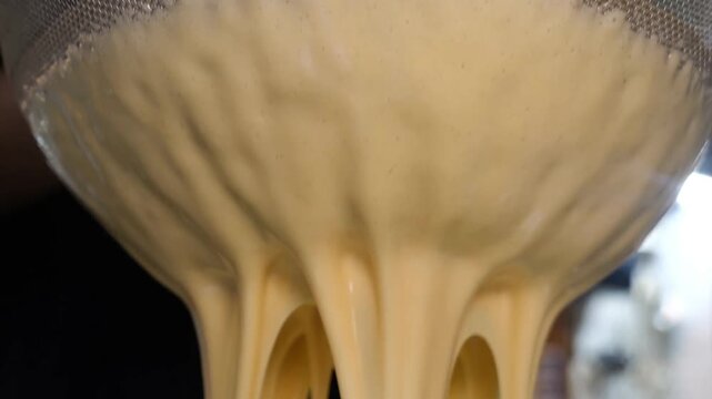A chef pours a large quantity of thick, smooth yellow batter from a bowl through a fine mesh strainer into a clear container, using a spatula to press the batter and ensure a consistent
