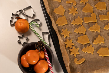 Unbaked gingerbread cookies rest on a parchment-lined baking sheet with metal cookie cutters, a bowl of mandarins, and candy canes. Ready to be baked for Christmas.