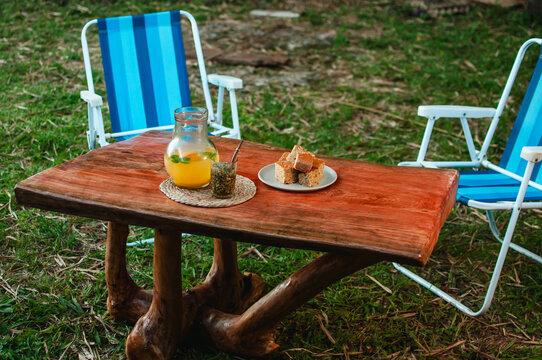 Un terer&eacute; de jugo de frutas con bocadillos sobre una mesa r&uacute;stica y silletas azules en el c&eacute;sped, momento de descanso al aire libre.